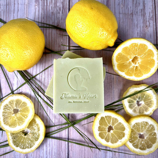A light green colored bar of soap with the words Mama's Heart All Natural Soap stamped on it surrounded by sprigs of green grass, whole lemons and lemon slices